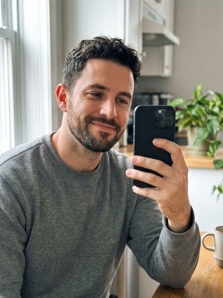 Original selfie: a man in a gray sweater holding his phone in a kitchen